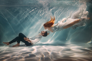 Happy couple in love swim underwater, female goddess muse inspires male writer poet creator. Nymph girl saves drowning guy at bottom sea under water. Red hair white long silk dress float. Art photo