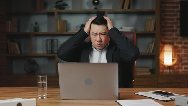 Front view of overwhelmed asian chief executive reading horrible news in internet while sitting at office desktop with portable computer. Perplexed man feeling shock and catching head with hands.
