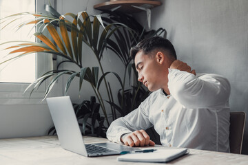 Exhausted young Caucasian male worker sit at desk massage neck suffer from strain spasm muscles. Tired unwell man overwhelmed with computer work sedentary lifestyle struggle with back pain or ache.. - Powered by Adobe
