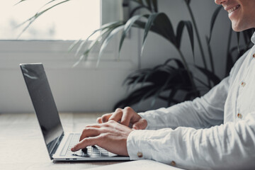Side and close view handsome young businessman working with computer remotely, sitting at wooden table in office. Portrait and close up hands writing and typing on laptop working..