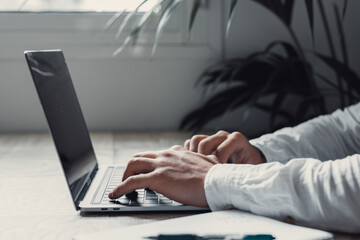 Side and close view handsome young businessman working with computer remotely, sitting at wooden table in office. Portrait and close up hands writing and typing on laptop working..