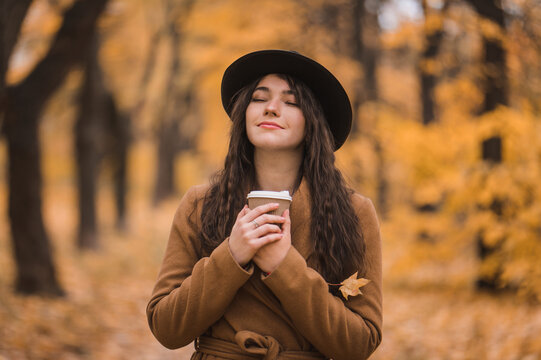 Cheerful young woman in beige coat and hat enjoys coffee from disposable cup in park in autumn. Girl drinks cappuccino on the street. walking in autumn park, happy mood, fashion style trend