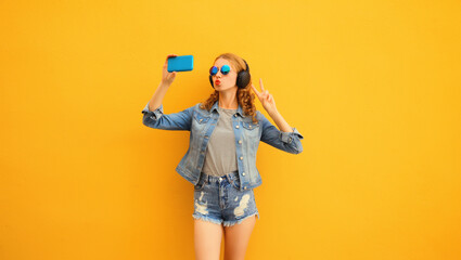 Portrait of happy young woman taking selfie with phone listening to music in wireless headphones wearing jacket on vivid yellow background