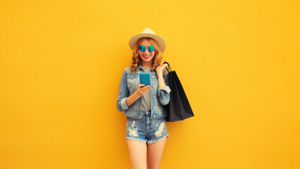 Beautiful happy young woman looking at phone with shopping bag wearing summer straw hat, denim jacket on yellow background