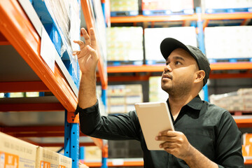 Warehouse worker wearing a hat and black shirt hands holding tablet check stock on tall shelves in...