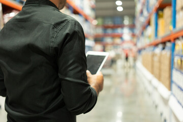 Warehouse worker wearing a hat and black shirt hands holding tablet check stock on tall shelves in warehouse storage. Asian auditor or staff work looking up stocktaking inventory in warehouse store.