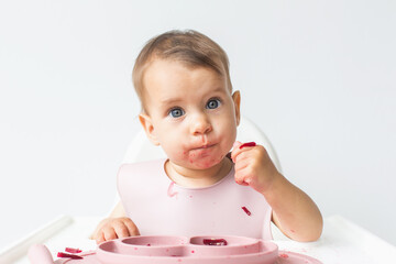 close-up portrait of a cute little girl 9 months old sitting in a highchair in the kitchen, looking at the camera, eating complementary foods