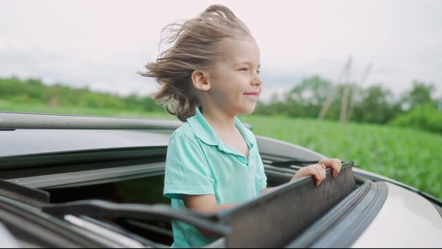 Adorable happy little boy stands in open car sunroof during road trip in countryside at summer. Concept of family leisure, active traveling. High quality 4k