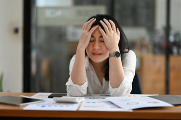 Portrait image of Young Asian businesswoman bowing and holding her head stressed expression with financial documents on working table.