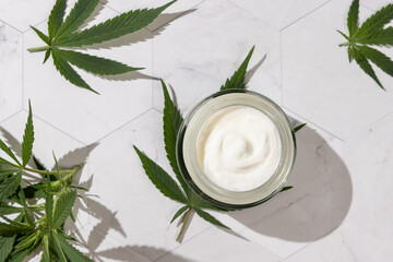 Opened cream jar near green cannabis leaves on white table top view, hard shadows