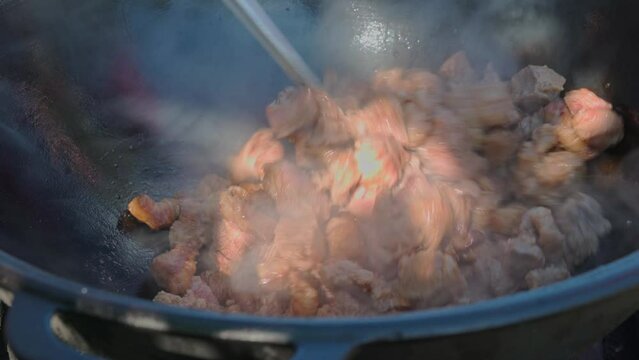 The cook fries lamb and stirs the meat with a spatula in cauldron close-up. Chef hand frying pieces of meat in a cast-iron pan for pilaf or bograch outdoor. Cooking on a bright sunny day in 4K 25FPS