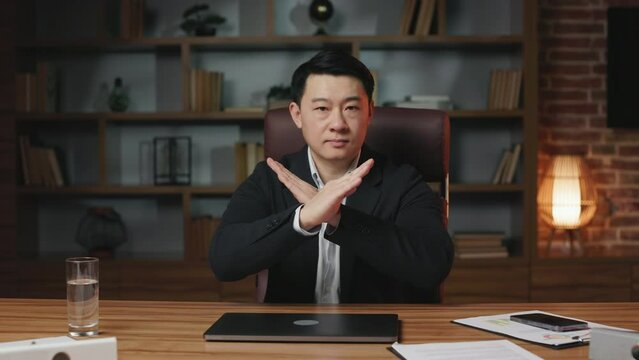 Front view of strict chief executive closing personal computer, crossing hands and saying no in trendy workplace. Serious asian boss dressed in formal outfit reaching palm forward and exclaiming stop.