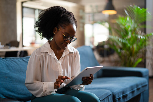 Beautiful African businesswoman sitting on sofa in office. Young woman using digital tablet.