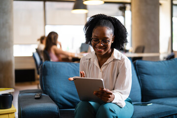 Beautiful African businesswoman sitting on sofa in office. Young woman using digital tablet.