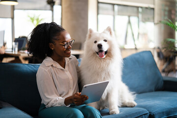 Beautiful African businesswoman sitting on sofa in office enjoying with dog. Young woman having video call.