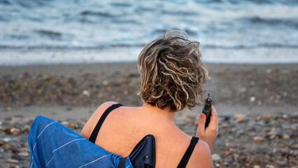 woman smoking electronic cigarette sitting on beach chair with her back to camera on the beach