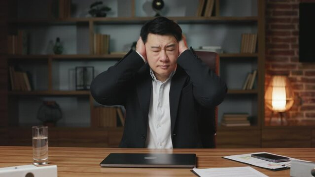 Portrait of stressed asian businessman closing ears with hands pretending not to hear anything. Troubled middle aged worker not willing to listen to silly advice and ignoring colleagues in office.