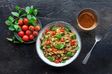 Quinoa tabbouleh salad in a bowl, a healthy dinner with tomatoes and mint, with a drink, overhead flat lay shot on a black slate background