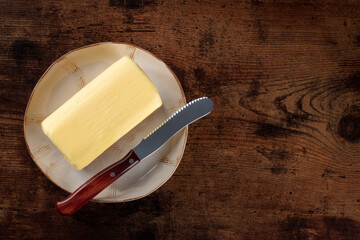 A block of butter with a knife on a plate, shot from above a dark rustic wooden background with...