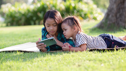 Fototapeta premium Two young girls lying on the lawn looking at their phones, summer, golden hour, sunset. SSTKHome