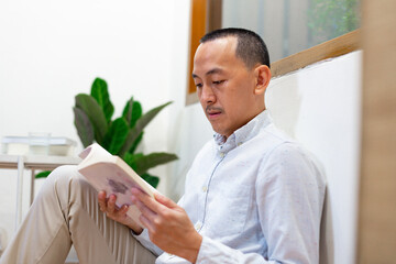 Stay home pastimes. Young man sitting in cozy armchair with open book indoors. Beautiful young man reading story, enjoying lazy morning, having relaxing weekend.