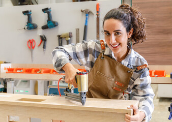Portrait Young Latin woman carpenter using nailer shooting on wood. to restore furniture in the...