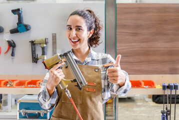Portrait young latino carpenter holding nailer looking at camera smiling in furniture carpentry workshop