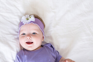 A baby with blue eyes wearing a purple outfit smiling lying on bed. top view