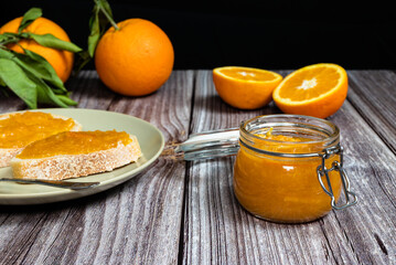 Delicious homemade orange marmalade in a glass jar. Jam on slice of bread on a plate. Horizontal photo with black background.