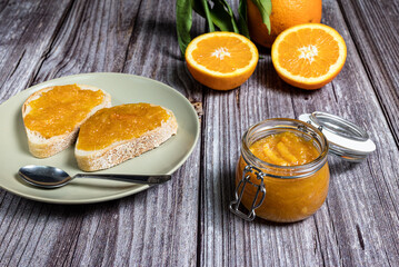 Delicious homemade orange marmalade in a glass jar. In the background slices of bread with marmalade in a plate on a white wooden table.