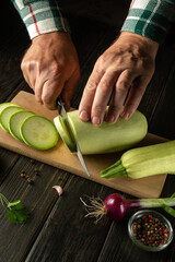 Hands of a chef with a knife cutting vegetable marrow or zucchini on a wooden cutting board before preparing vegetarian food