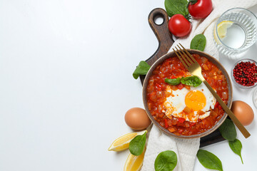 Shakshuka in bowl on board, fork, towel and ingredients on white background, space for text