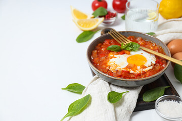 Shakshuka in bowl on board, fork, towel and ingredients on white background, space for text