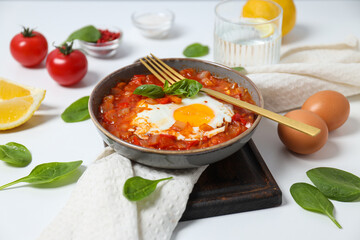 Shakshuka in bowl on board, fork, towel and ingredients on white background