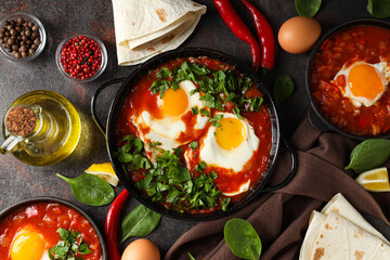 Pans with shakshuka, eggs, basil, bowl of pepper and oil on dark background, top view