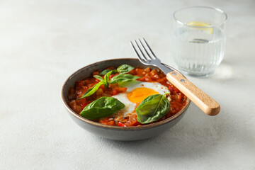 Shakshuka in bowl, fork and glass of water on light background
