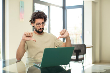 young adult bearded man with a laptop looking sad, disappointed or angry, showing thumbs down in disagreement, feeling frustrated