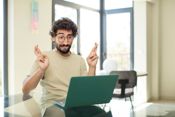 young adult bearded man with a laptop crossing fingers anxiously and hoping for good luck with a...