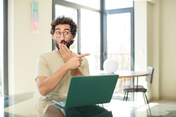 young adult bearded man with a laptop feeling surprised with a shocked expression and pointing to the side