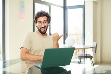 young adult bearded man with a laptop smiling cheerfully and casually pointing to copy space on the side, feeling happy and satisfied