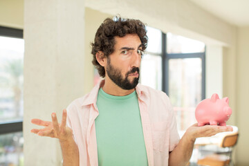 crazy bearded man shrugging, feeling confused and uncertain. piggy bank concept
