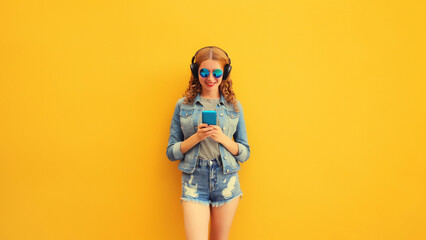 Portrait of stylish modern happy young woman listening to music in headphones with phone wearing jacket, sunglasses on yellow background