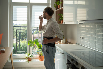 Lifestyle and spinal disc herniation. Man wearing lumbar brace drinking water while standing in kitchen at home, using lumbar corset to treat low back pain, daily living after spinal injury or surgery