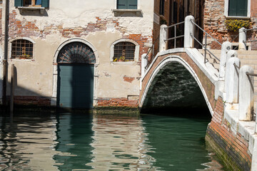 Shabby old walls over the water of a canal in Venice. A fragment of a building and a bridge with crumbling plaster, green shutters on windows and arched door.