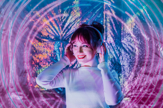 Portrait of relaxed smiling woman in white clothes and headphones dancing in neon light. Music lover. White dress code party. Silent disco. Enjoy moment on shiny background. Selective focus.