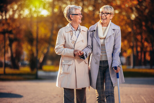 An older woman helping another woman that is walking with a cane in the park in the autumn