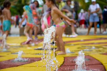 Little happy children in bright swimsuits run and jump over a stream of water gushing from a...