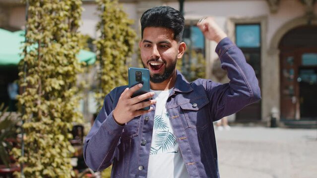 Young bearded indian man use mobile smartphone celebrating win good message news, lottery jackpot victory, giveaway online. Happy hindu guy walking outdoors in urban city street background. Lifestyles