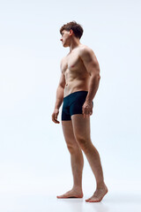 Full-length image of young man with muscular body posing shirtless in underwear against grey studio...