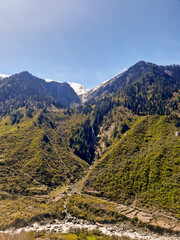 A beautiful view of waterfall in Sankari Range from a distance with a green grassy field and huge pine trees on a sunny summer day. A view from har ki dun trek
